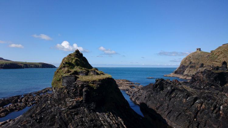 Jagged angular outcrops of slate rock set against a blue sky with the sea and coast behind