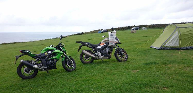 The 2 motorcycles and the tent set against the sloping field and the hazy sea at Caerfai Bay