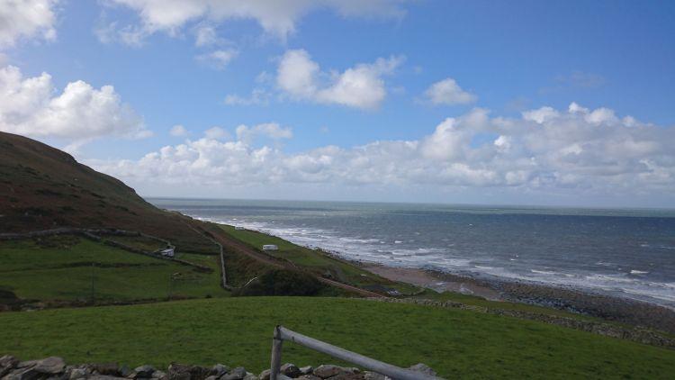 Hills roll down across fields to a narrow shoreline then the sea. Blue skies with light clouds