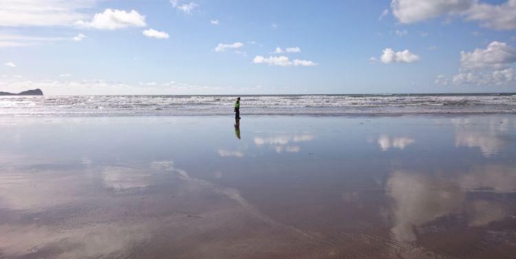 Ren stands alone at the shoreline with the sky reflected off the wet sands of the beach