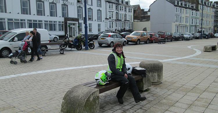 Sharon sat on a bench smiling with a large white seafront hotel behind her