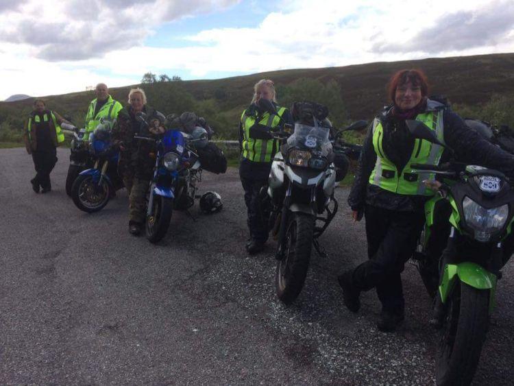 5 riders stand by their machines on a Highland road, all smiling at the camera