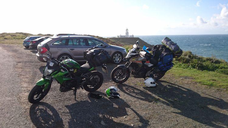 Strumble Head lighthouse is in the distance with the motorcycles and some parked cars in front