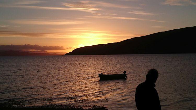 A deep orange sunset, a loch, a small boat and a person in outline at Ullapool
