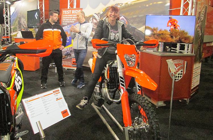 Sharon is sat on a very tall KTM off road bike at a bike show. Her feet are about 1 foot from the floor