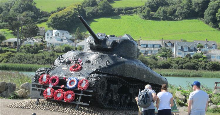 A very damaged and painted in thick black paint Sherman tank. There are wreaths and memorials on it
