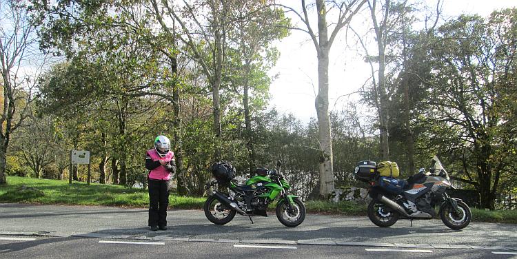 Sharon is looking at her phone next to the motorcycles amidst trees and gorgeous sunshine
