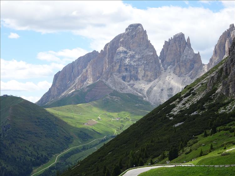 A road winds between steep green hillside and enormous rocky mountain tops in the Dolomites