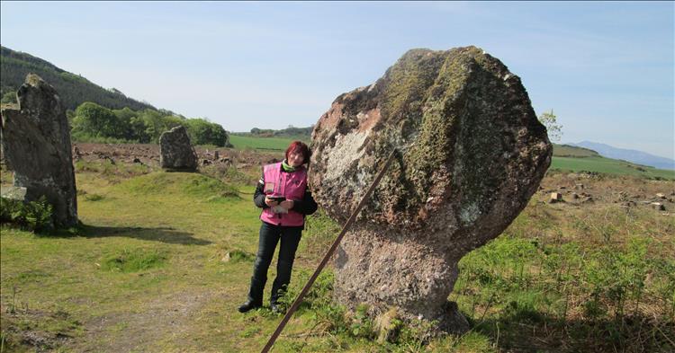 Sharon leans against one of the 3 standing stones on open ground with tree stumps all around