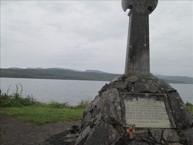 A stone cross and plaque with misty moist hills across the calm flat Holy Loch