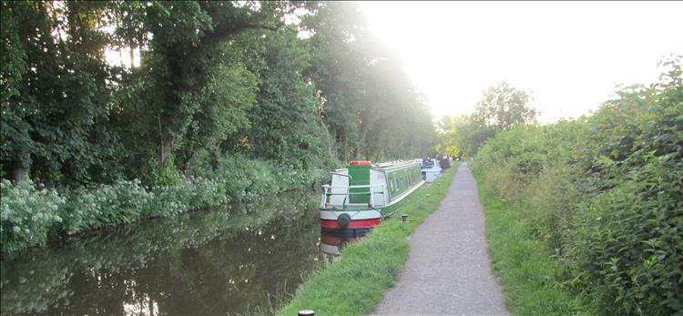 The towpath and canal with a boat moored up all set among trees and the setting sun