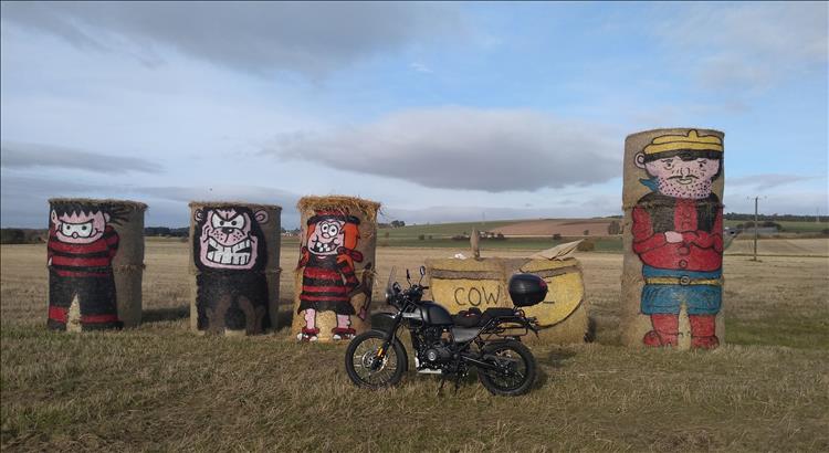 The Royal Enfield Himalayan in a field with large straw bales painted to look like Beano Characters