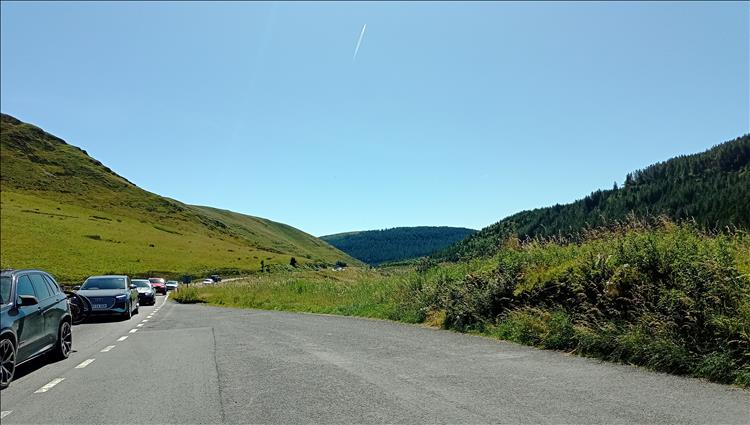 With a few cars in a queue the scene is of hills and trees and grasses and blue skies