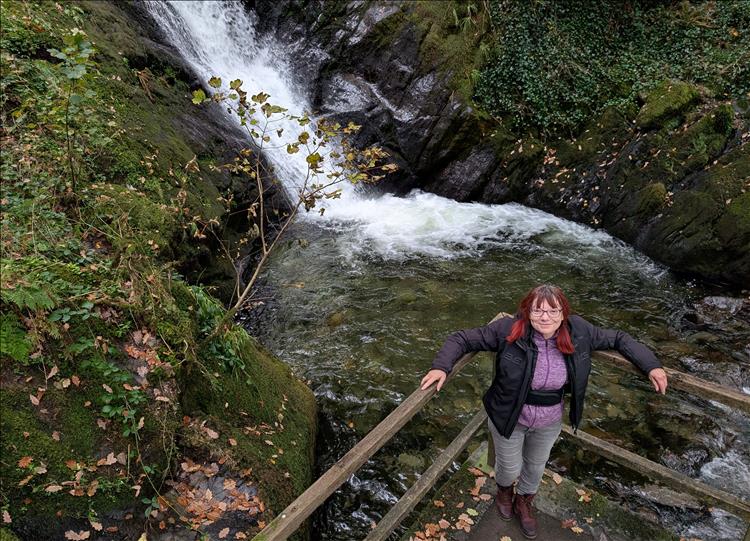 Sharon poses by a narrow but fast waterfall with rocks and trees