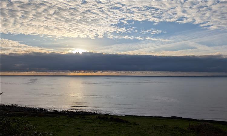 A dead calm sea, thick clouds and light clouds with the sun just poking from behind a cloud