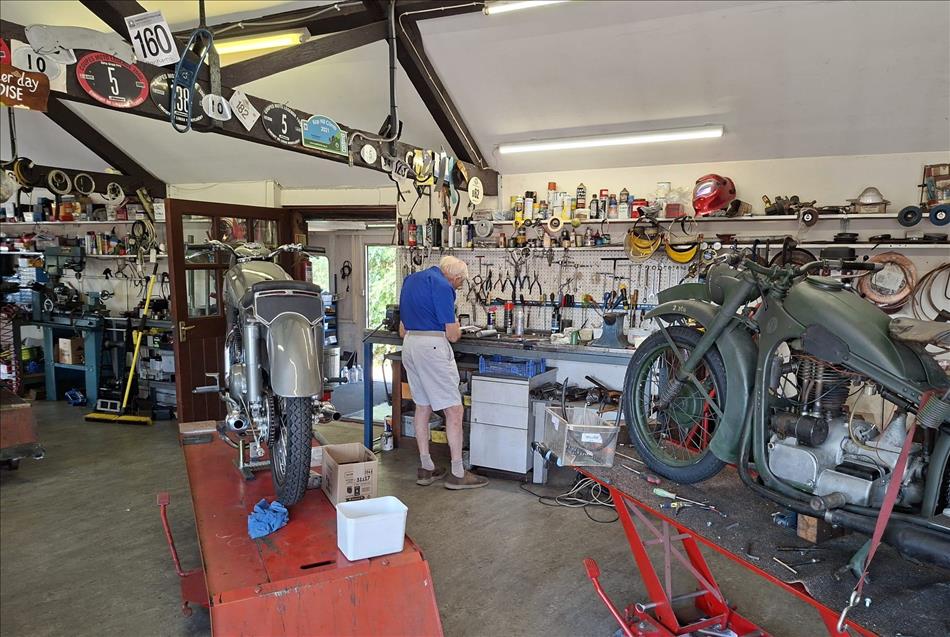 A workshop with old motorcycles on lifts, trinckets everywhere and an old chap working at a bench