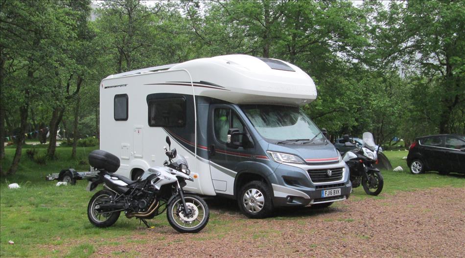 A motorhome with a trailer and 2 motorcycles beside the motorhome on a campsite in the trees