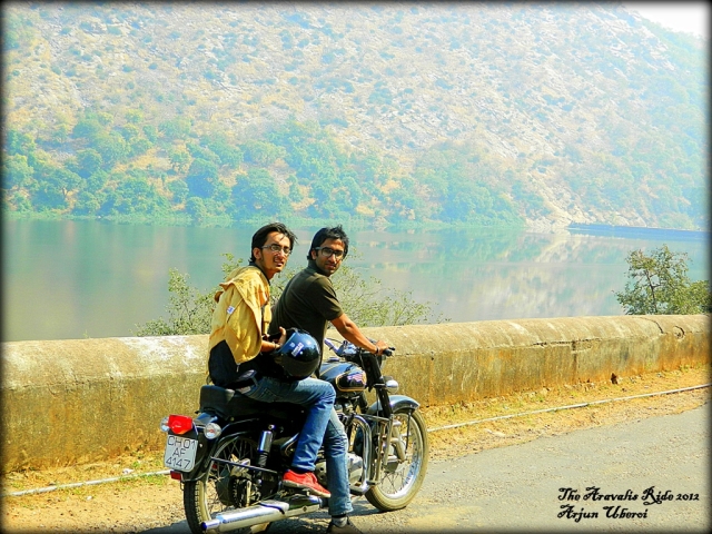 2 slim young indian men sat on an enfield set by a lake and hills