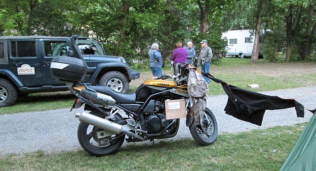 my motorcycle next to the tent with clothes drying on a cord between them. big 4 by 4 behind and people talking