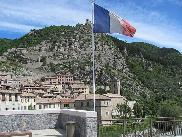 a rocky hillside with a town clinging to the slopes, a french flag in the foreground