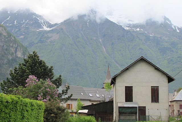 alpine house with steep jagged mountains behind, topped with mist. The road to le bourg de oisans