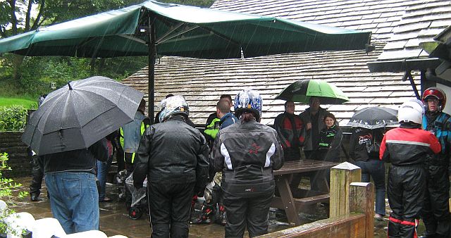 a group of motorcyclists shelter from the rain under a canopy and umbrellas