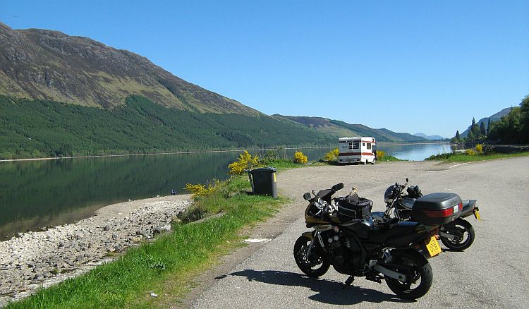 a still calm scottish loch in the glories sun and clear skies with 2 motorcycles in the foreground