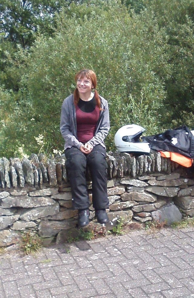 sharon sits on a stone wall i her bike gear smiling in the welsh sun