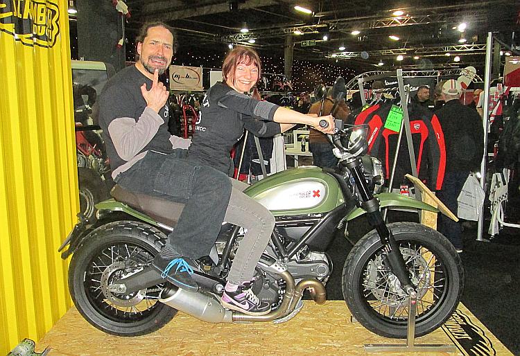 Sharon and Ren sat on a Ducati at a bike Show. Ren is on the back and looks scared
