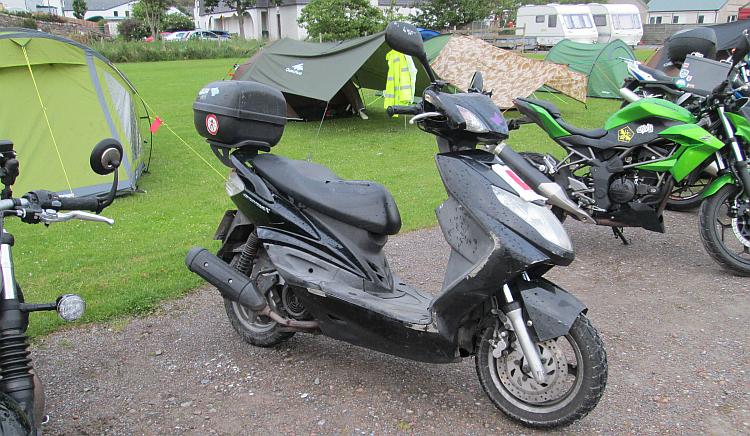 A scooter with learner plates on a campsite with other motorcycles