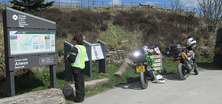 Sharon stands beside the bikes and the tourist signs at Alwen Reservoir