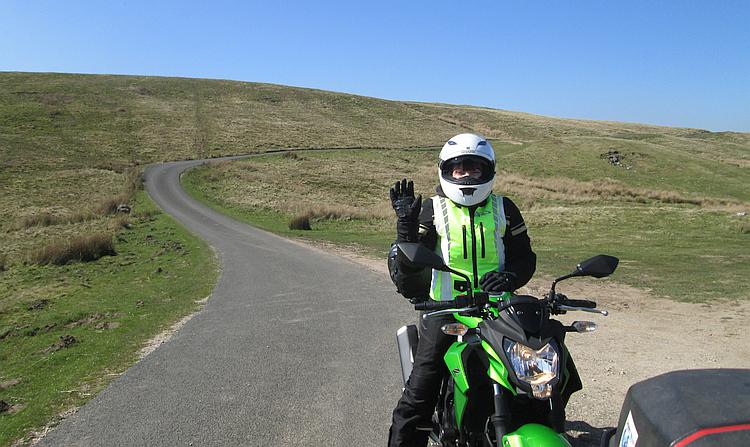 Sharon sits on her bike in all her gear waving at the camera surrounded by beautiful folling hills