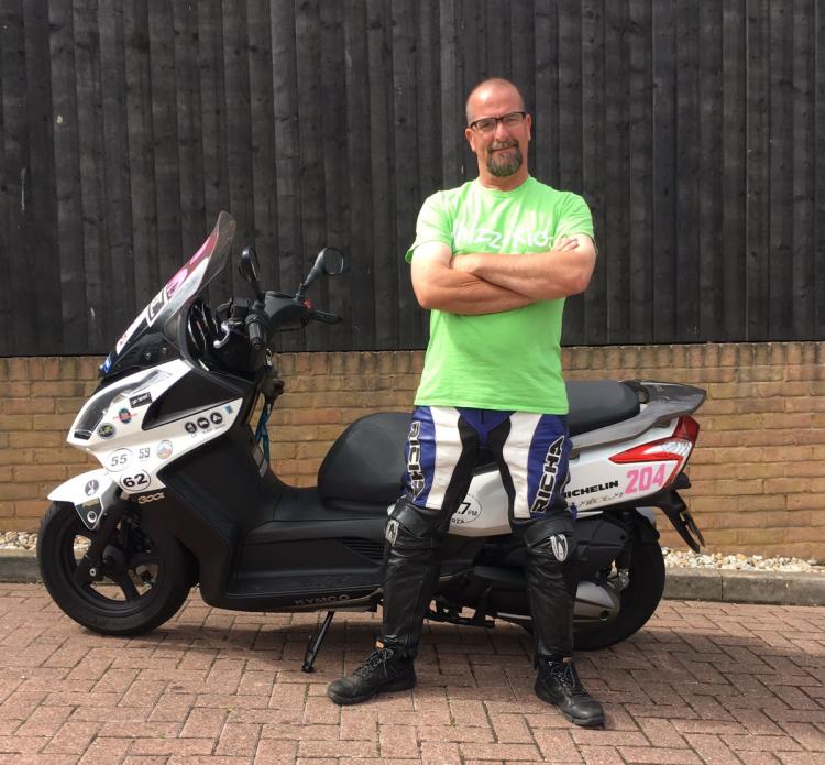 Stephen, a strong bald man in leathers, stands beside his 300cc Kymco scooter