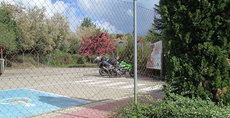Behind a wire fence the bikes are in a car park surrounded by trees at the site