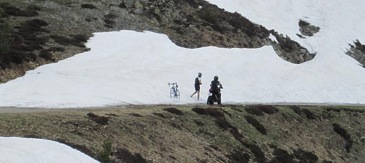 Against the snow atop a mountain a cyclist and motorcyclist talk in the distance
