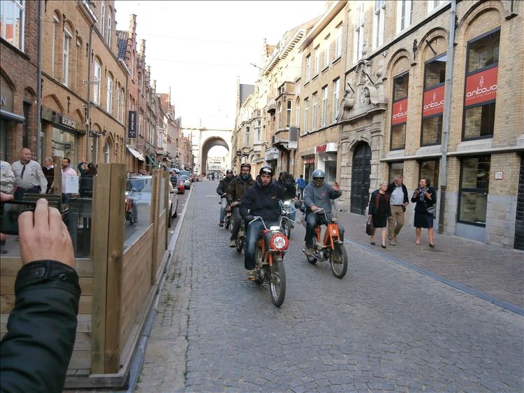 About 6 or 7 riders all on very small Honda scoots riding down the streets of Ypres
