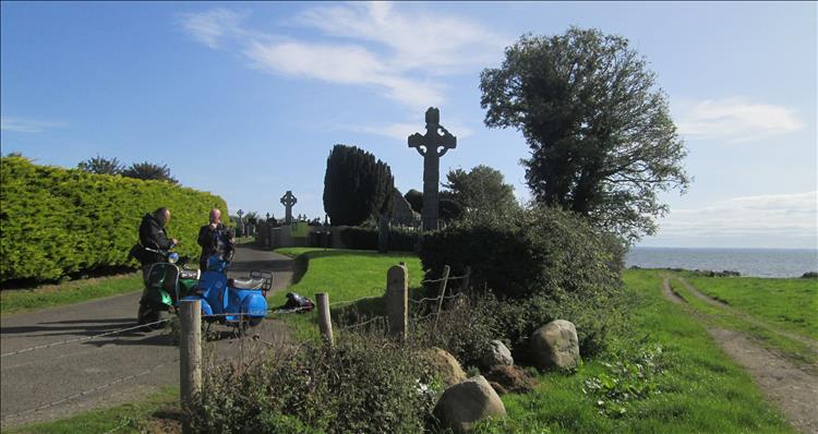 An ancient tall stone cross in the countryside with 2 scooters in the foreground