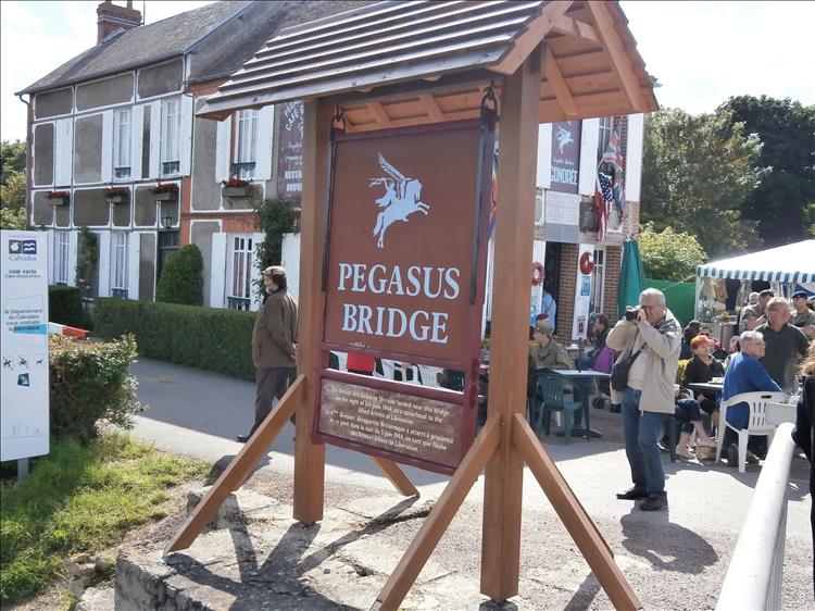 A wooden sign stating Pegasus Bridge outside a french building, bust with tourists