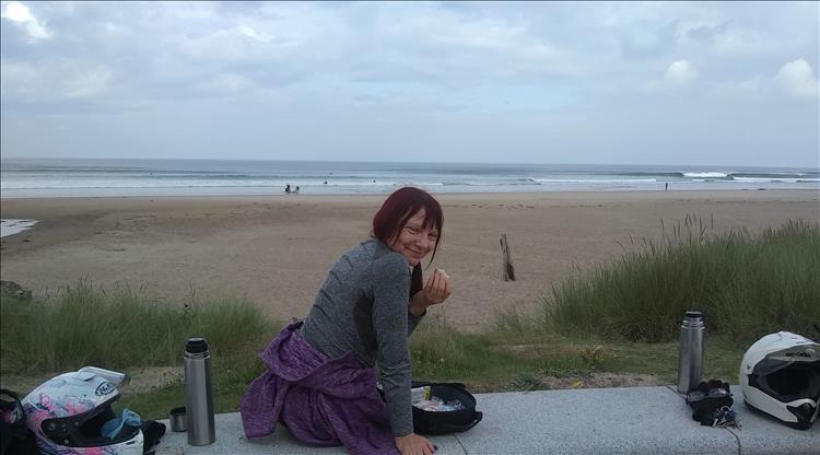 Sharon's happy eating with a big sandy beach and grey skies behind her