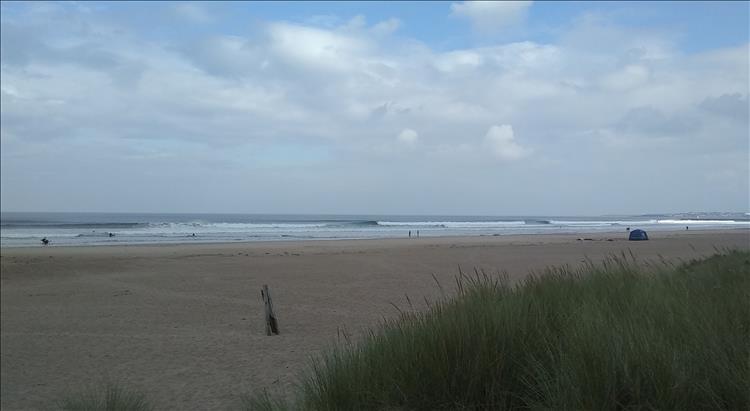 A vast sandy beach, large waves and in the distance people enjoy the surf