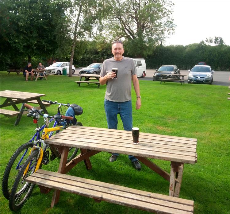 A friendly face smiles while holding a pint, 2 old mountain bikes are propped against the bench