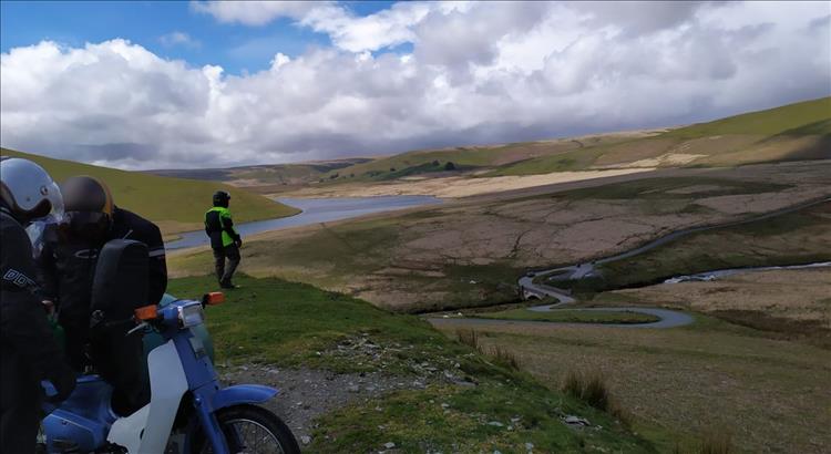 2 chaps looking at a Honda Cub while another admires the glorious vast hills aand lakes in Wales