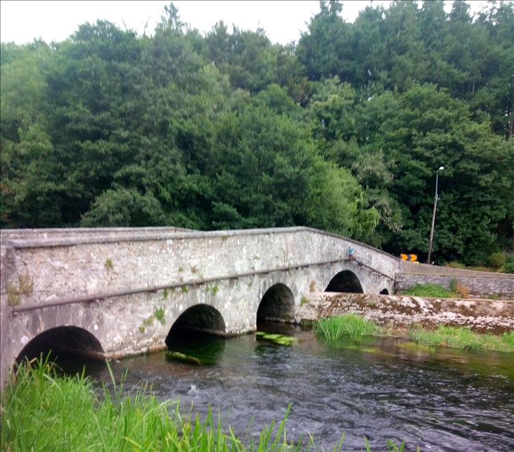 A low old stone bridge crosses a river among trees and grasses