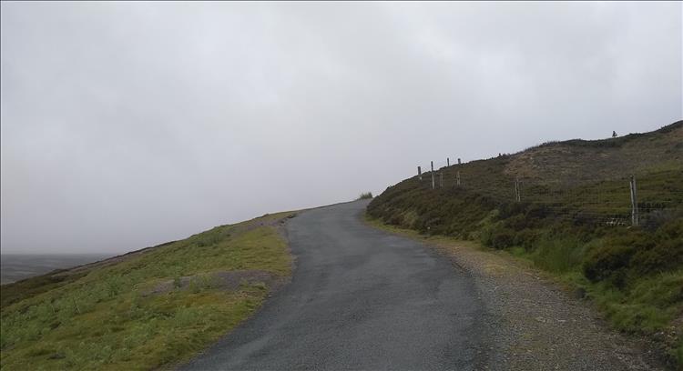 A steep single track lane rises up and wriggles up the side of a hill, grey skies all around