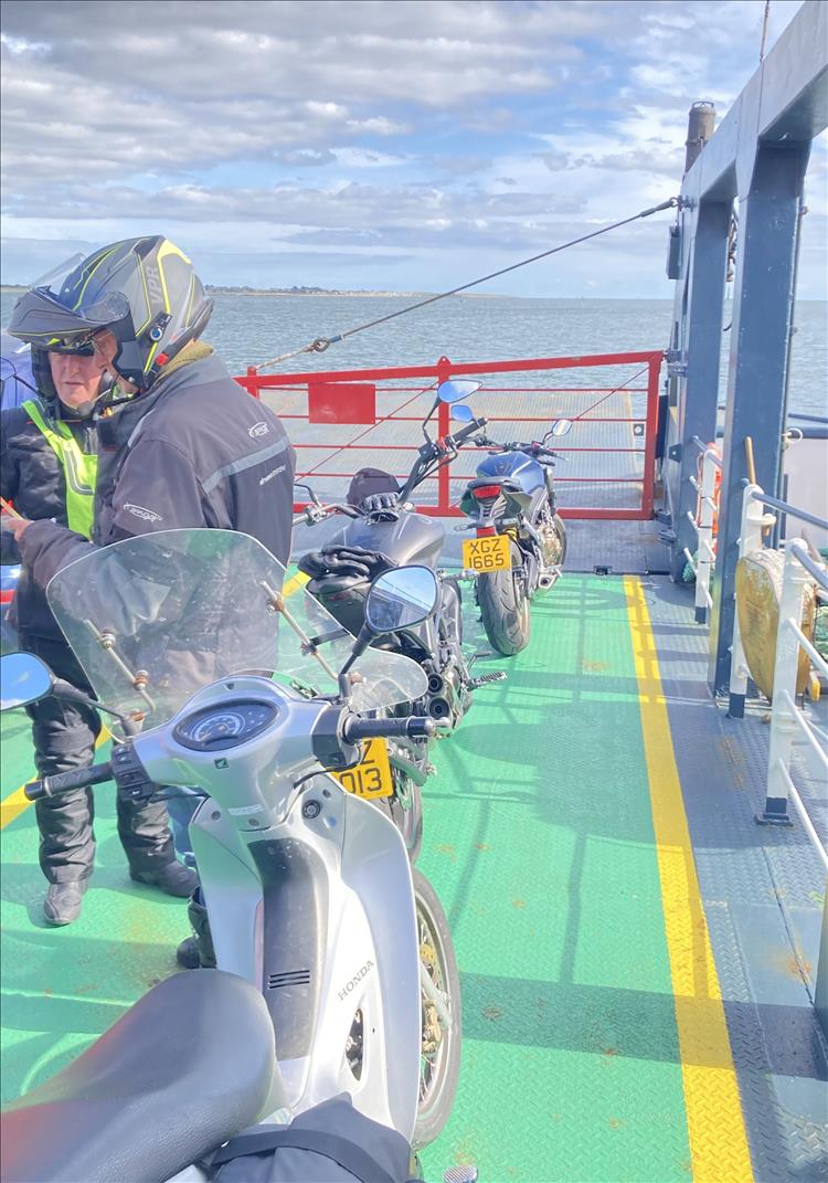 The lads talk in their bike gear stood next to the bikes and the ferry crosses the calm waters
