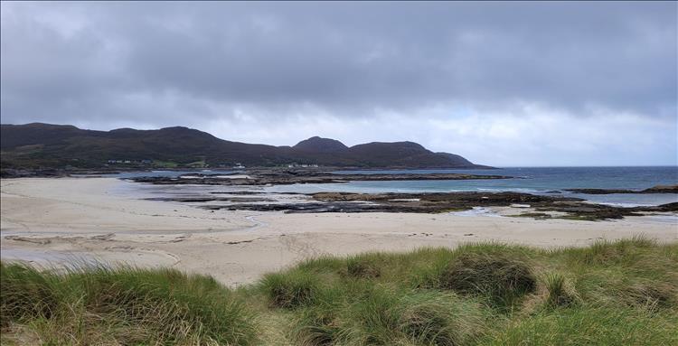 A broad sandy beach with low rocks amid the sand and sea, big rugged hills in the background at Sanna