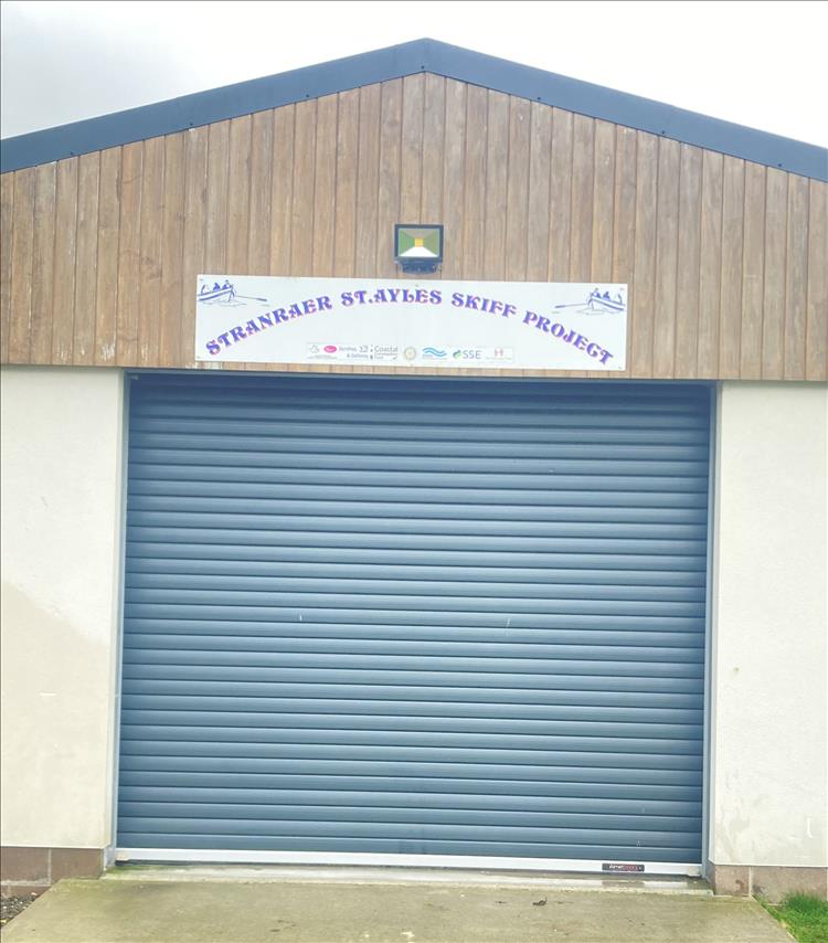 A large roller shutter in a large shed, the sign above reads Stranraer St.Ayles Skiff Project
