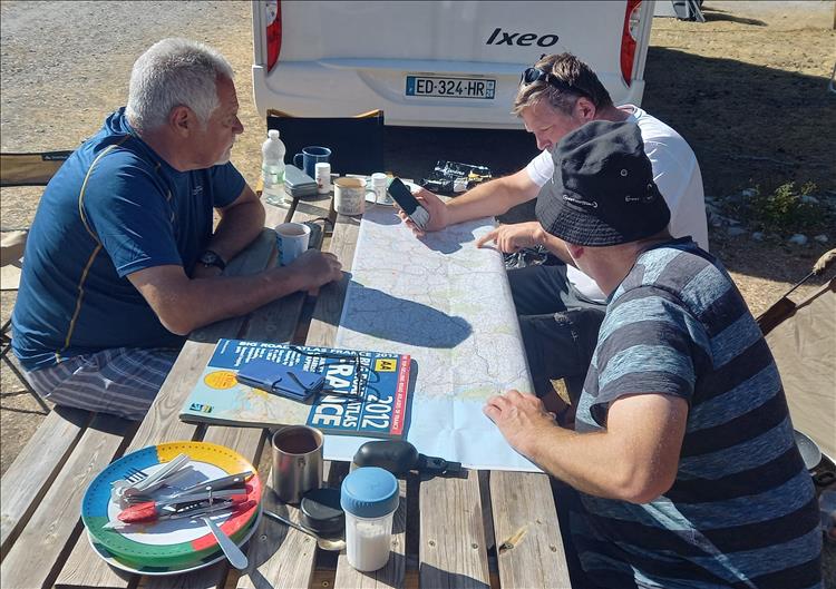 Three older chaps sit around a bench table looking at maps in the morning sun