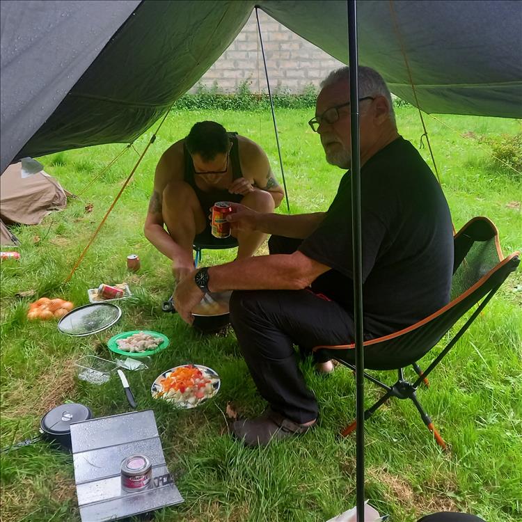 2 chaps cooking and drinking under a tarp on grass