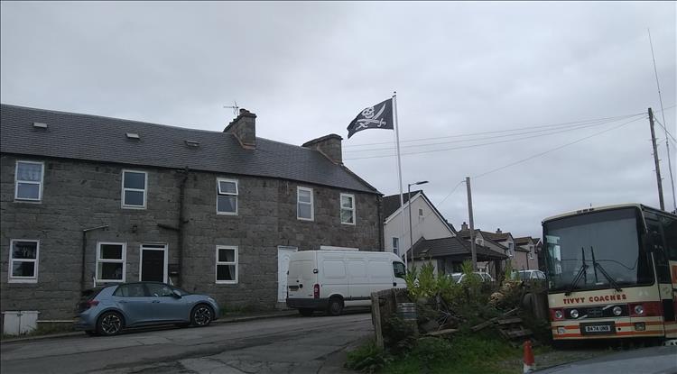 A Jolly Roger flag atop a pole beside a scruffy workshop